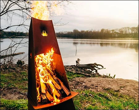 A tall, conical wood-burning fire pit with flames burning inside. The fire pit is surrounded by green grass and is near a body of water. There is a lake in the background.