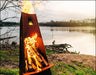 A tall, conical wood-burning fire pit with flames burning inside. The fire pit is surrounded by green grass and is near a body of water. There is a lake in the background.