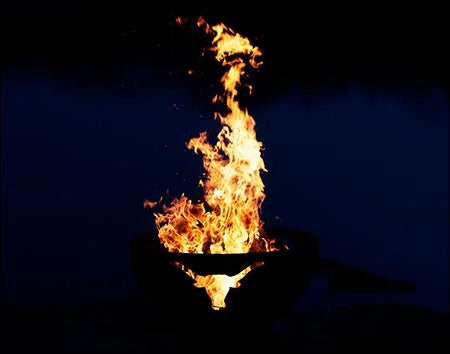 A close-up image of a wood-burning fire pit with a longhorn cutout design. The fire is burning brightly, illuminating the cutout. The background is dark.