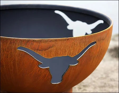 A close-up image of a wood-burning fire pit with a longhorn cutout design. The cutout is painted white against a rusty background.