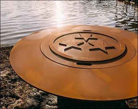 A close-up of a round, rust-colored fire pit lid with a starburst design in the center. The lid is placed on a rock near a body of water.