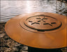 A close-up of a round, rust-colored fire pit lid with a starburst design in the center. The lid is placed on a rock near a body of water.