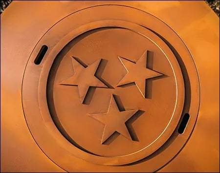 A close-up of a round, rust-colored fire pit lid with three stars in the center. The stars are arranged in a triangular pattern.