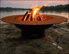 A round, rust-colored wood-burning fire pit with flames burning inside. It is placed on a rock near a body of water with a sunset in the background. There are two champagne glasses on the ground next to the fire pit.