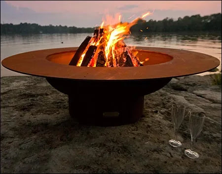 A round, rust-colored wood-burning fire pit with flames burning inside. It is placed on a rock near a body of water with a sunset in the background. There are two champagne glasses on the ground next to the fire pit.