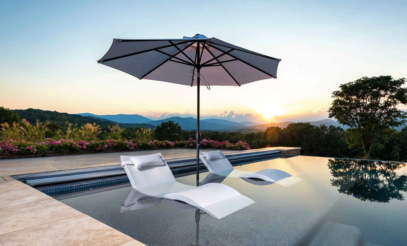 A photo showcasing two white floating lounge chairs with plush pillows, offering a relaxing spot to enjoy the sunset. The pool overlooks a picturesque landscape with rolling hills and a vibrant sky. A large umbrella provides shade from the setting sun.