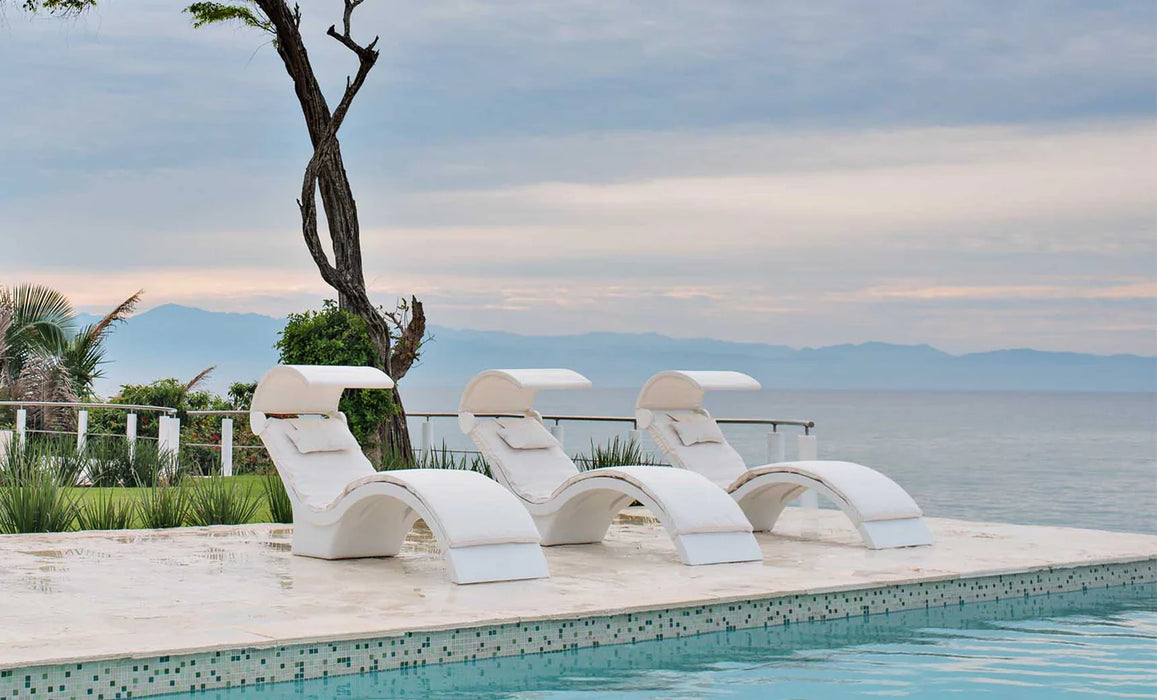 Three white, curved pool loungers, likely Ledge Lounger Signature Chaise Deep or similar, are positioned in a row on a light-colored tiled patio overlooking a calm ocean bay. Each lounger features a retractable white sun shade and plush white cushions. 