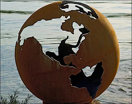 A spherical  wood burning fire pit with a map of the Earth etched into its surface. The fire pit is made of a rusted metal and is placed on a rock near a body of water. The continents of North America, South America, Europe, Africa, Asia, Australia, and Antarctica are visible on the surface of the fire pit.