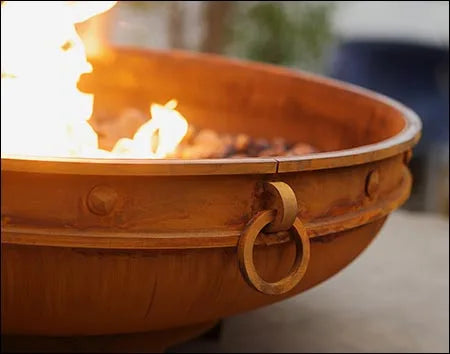 A close-up of a round, rusted metal fire pit with flames burning inside. The fire pit has two handles on the side and a raised edge.