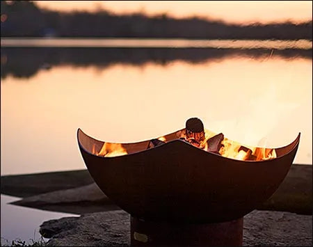  A manta ray-shaped fire pit with flames burning inside. The fire pit is placed on a rock near a lake at sunset.