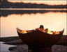  A manta ray-shaped fire pit with flames burning inside. The fire pit is placed on a rock near a lake at sunset.