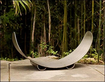 An empty carbon steel crescent firewood log rack placed in a garden setting with bamboo plants in the background.