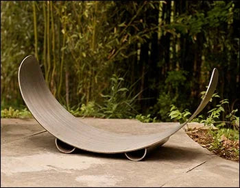 An empty carbon steel crescent log rack placed in a garden setting with bamboo plants in the background.