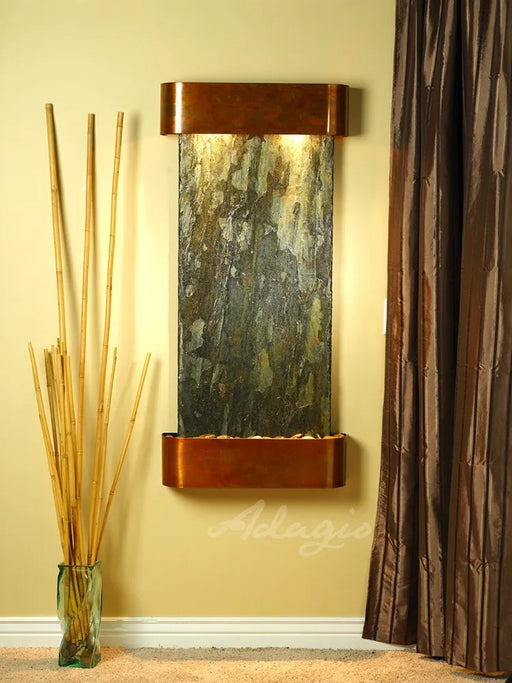 
A close-up, vertical shot of an Adagio Cascade Springs wall-mounted water fountain. It features a rich, antique copper frame with a strongly textured, multi-toned green and grey natural slate face. Warm lights from the top illuminate the water cascading down the slate into a lower basin filled with light-colored river rocks.