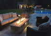 A wooden bench with a slatted back and seat sits on a stone patio. A round wooden table with a pedestal base complements the bench.