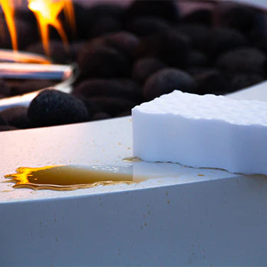 Detail shot of cleaning a white fire pit surface with a sponge, showing a dark spill being wiped away, with a flame and rocks in the background.