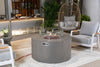 Light gray Elementi Pacaya 43.3" Round Hidden Propane Tank Fire Pit Table with flames and a glass wind guard, set on a patio with gray armchairs, a hanging chair, and plants.
