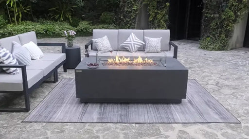 dark grey luxury fire table has a fire lit in the middle. fire table is placed on top of a rug in the middle of sofas