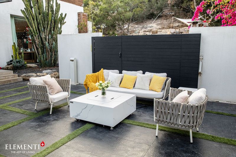 fire table used as a table to hold a vase. it is white marble with black accents. it is placed in a beautiful patio surrounded by pretty flowers. the fire table is placed in the middle of comfy seats