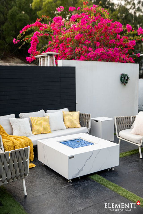 white fire table that is square and marble with black accents. In the fire tables pit there is blue fiber glass laid out but not lighting the fire. The fire table is placed in the middle of 2 single seats facing each outer and 1 three seater seat in a patio