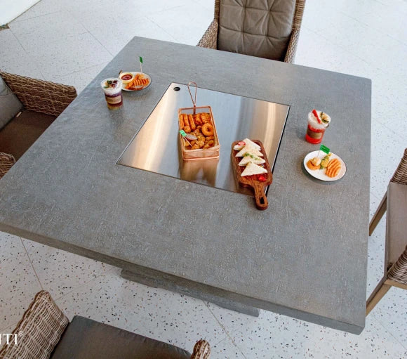 A square outdoor dining table made of concrete with a stainless steel fire pit insert. The table is surrounded by wicker chairs and has food and drinks placed on it.