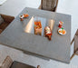 A square outdoor dining table made of concrete with a stainless steel fire pit insert. The table is surrounded by wicker chairs and has food and drinks placed on it.