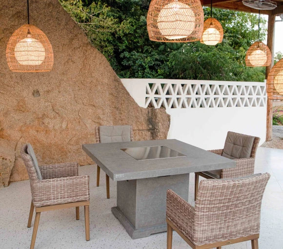 A square outdoor dining table made of concrete with a stainless steel fire pit insert. The table is surrounded by wicker chairs and is situated on a patio with a rock wall and hanging lanterns in the background.