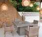 A square outdoor dining table made of concrete with a stainless steel fire pit insert. The table is surrounded by wicker chairs and is situated on a patio with a rock wall and hanging lanterns in the background.