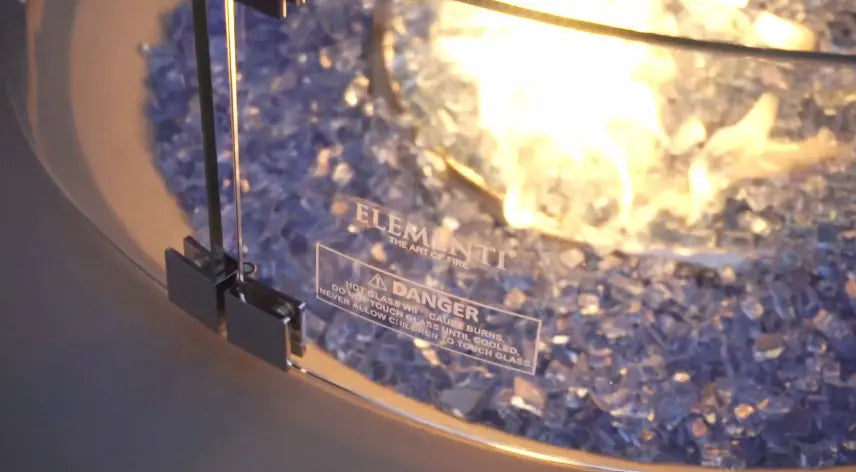 Close-up of a fire pit with flames, blue glass pieces, and a glass wind guard showing a warning label.