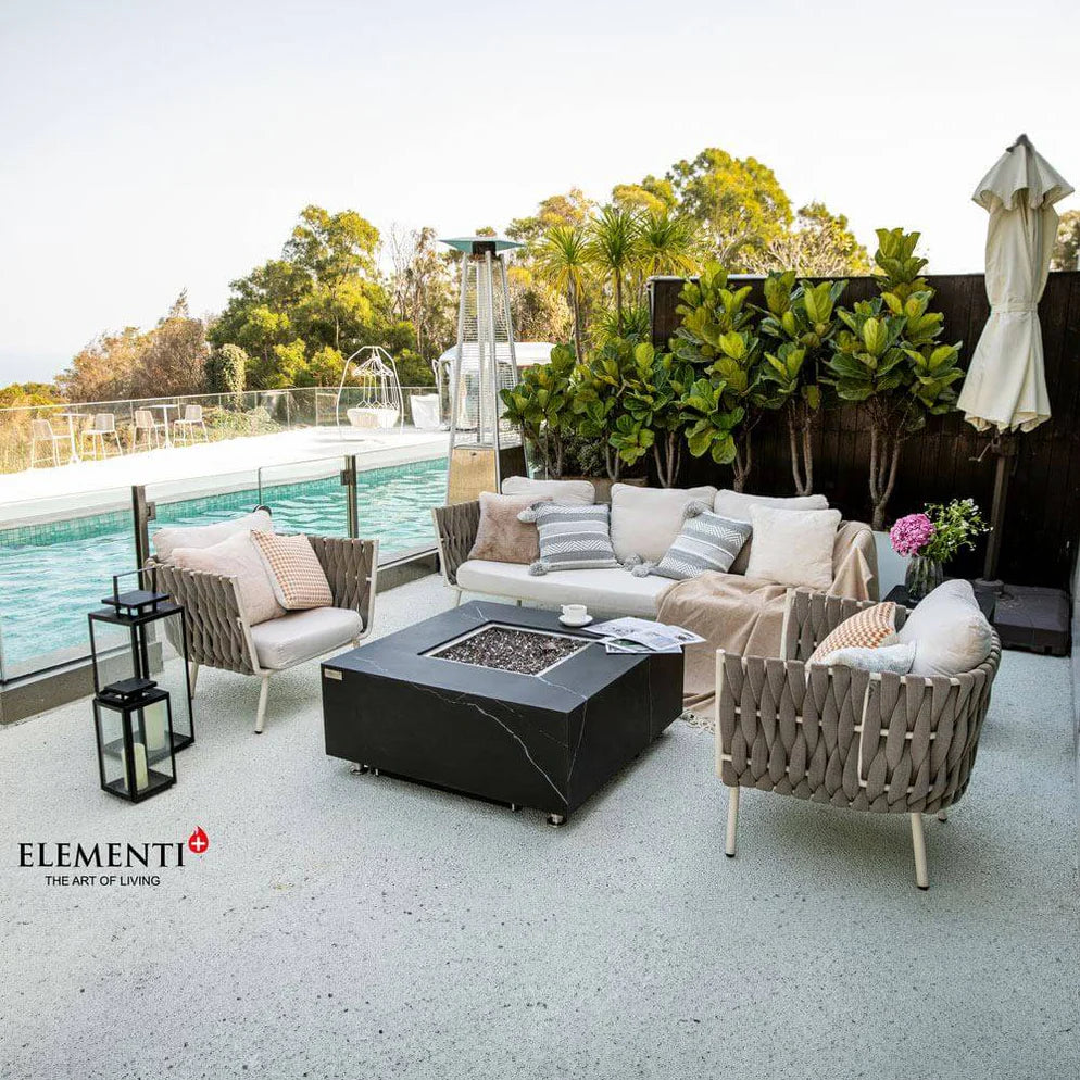 Modern outdoor patio featuring a square black fire pit table with a glass surround, surrounded by a white sofa and chairs with light cushions. The patio overlooks a swimming pool and lush greenery.