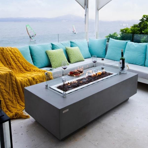 rectangular fire pit coffee table by elementi. fire pit table is lit with windshield protecting flame. There are wine glasses and bottle of wine on top of table in a beautiful outdoor setting