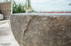 A close-up shot of the textured surface of a round Elementi fire table, with red lava rocks visible in the center. The "ELEMENTI THE ART OF LIVING" logo is in the bottom left corner.