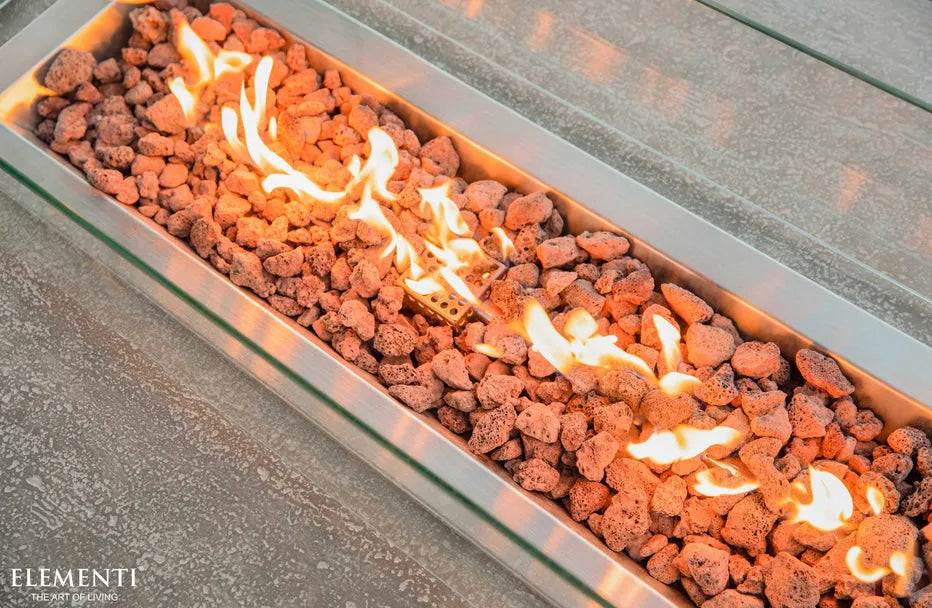  A close-up, angled view of a rectangular fire pit burner. Flames flicker and dance above a bed of dark red lava rocks, contained within a long, rectangular metal tray. The edge of a gray concrete fire pit table is visible at the top and bottom of the image.