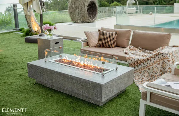 A rectangular concrete fire pit table with a glass wind guard and visible flames sits on a green patio overlooking a swimming pool. A beige sofa with pillows and a side table are in the background.