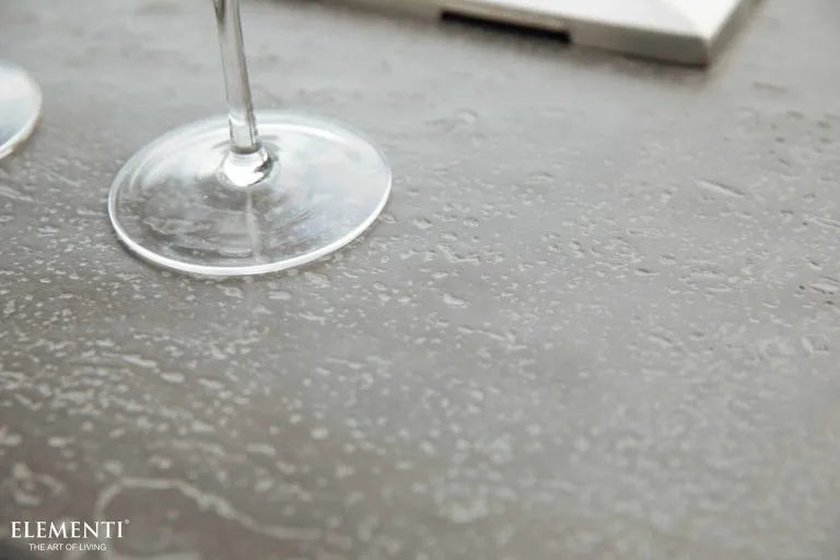 A close-up of a textured, gray surface with two wine glasses resting on it. The surface has a rough, almost concrete-like appearance with visible grains and imperfections. The word "ELEMENTI" is embossed in the lower right corner, with the phrase "THE ART OF LIVING" below it.