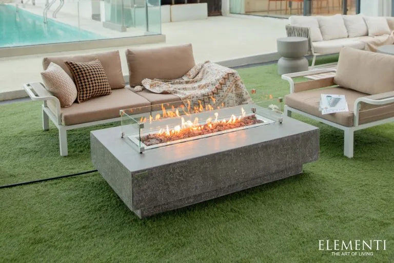 A rectangular concrete fire pit table with a glass wind guard. The fire pit is burning with a bright orange flame. The table is surrounded by a white sofa and two armchairs on a green lawn. A swimming pool can be seen in the background.