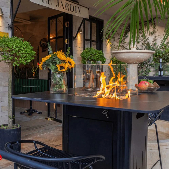 elementi fire pit table with glass windscreen. fire pit is lit with yellow and orange flames