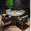A slightly elevated, full shot showcases a modern round dining table set for four in an indoor or semi-outdoor space. The table has a smooth, light grey top and a cylindrical pedestal base with horizontal striations.