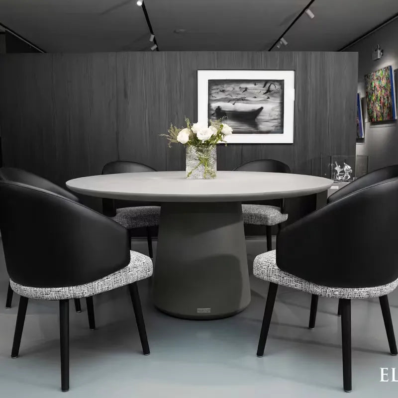 A round grey dining table with a pedestal base surrounded by six black and grey chairs in a modern setting.