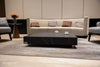 A square coffee table with a dark black marble porcelain top featuring white veining, and a matching black base with silver feet, sits in a living room. Two white cups are placed on the table.