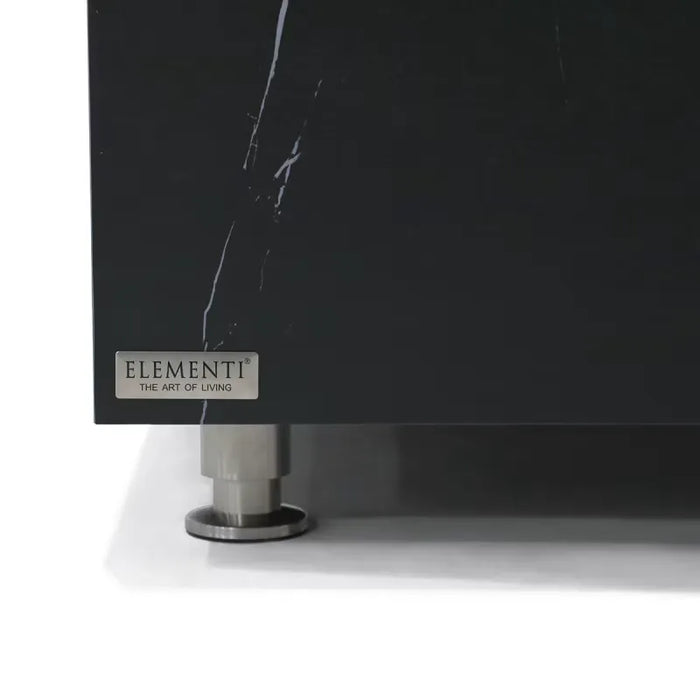A detailed view of the corner of a black marble porcelain table. The "ELEMENTI THE ART OF LIVING" logo is visible on a small plaque. The table rests on a short, cylindrical, silver leg with a wider, flat base.