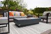 An eye-level, wide shot captures a stylish outdoor patio setting. The focal point is a substantial, dark gray rectangular coffee table with a textured, concrete-like surface and a slightly smaller base.