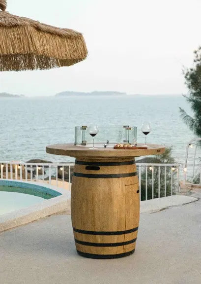 A round, wooden barrel fire table with a glass top. The table is placed on a patio overlooking a pool and ocean. There are wine glasses and a charcuterie board on the table.