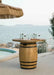 A round, wooden barrel fire table with a glass top. The table is placed on a patio overlooking a pool and ocean. There are wine glasses and a charcuterie board on the table.