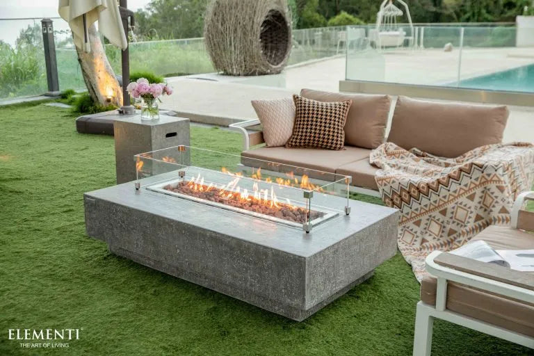 A rectangular concrete fire pit table with a glass wind guard. The fire pit is burning with a bright orange flame. The table is surrounded by a white sofa and two armchairs on a green lawn. A swimming pool can be seen in the background.