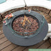 elementi dark grey round fire pit bowl with glasses and fruit platter on the side of the table. fire pit is lit with glass windshield around it