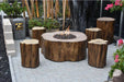 A round, wooden fire pit table surrounded by four wooden stools. The fire pit is burning with flames and has lava rocks inside. The table and stools are placed on a patio with bamboo fencing and plants in the background.