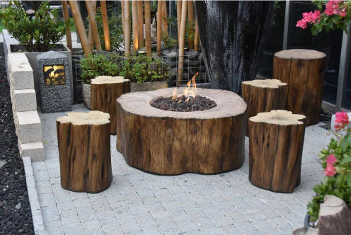 A round, wooden fire pit table surrounded by four wooden stools. The fire pit is burning with flames and has lava rocks inside. The table and stools are placed on a patio with bamboo fencing and plants in the background.