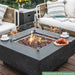 dark grey square fire pit table with fruit platter and coffee cups on table. fire pit is lit on brown lava rocks and square glass windscreen