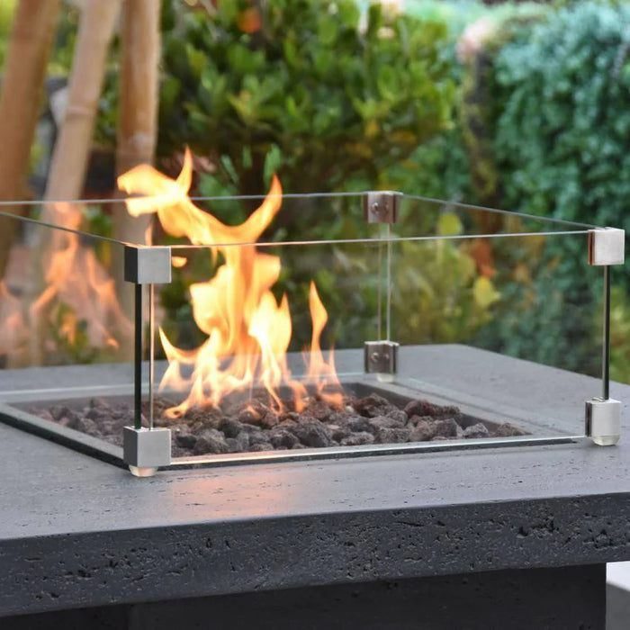 bar height fire pit table with square glass windscreen is protecting fire flame that is lit on lava rocks